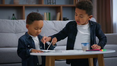 Two African American children painting at home enjoying paint together ethnic family sons boys kids siblings relatives in living room creating picture watercolor colors art hobby weekend entertainmentの写真素材