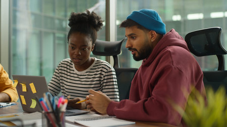 Arabian businessman talking with smiling African American businesswoman looking at laptop together...