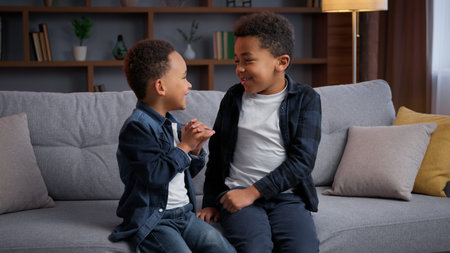Two little African American boys talking fighting playing at home on couch together communicate ethnic brothers children kids siblings sons talk fooling around having fun conversation cheerful familyの写真素材