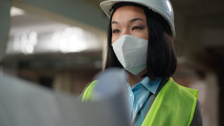 Asian Korean Chinese Japanese woman developer in safety mask hard hat female engineer builder architect inspector looking at building project plan construction girl checking blueprint industry workerの写真素材