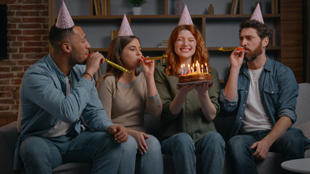 Four multiracial friends singing happy birthday song to Caucasian woman holding festive cake with candles smiling satisfied congratulate girl at home party diverse teammates women men blowing whistlesの写真素材