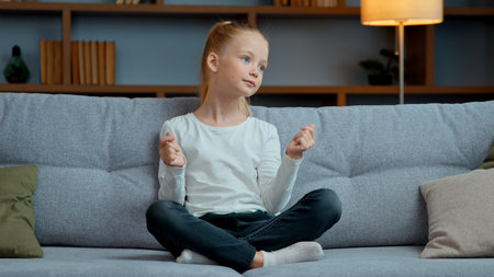 Little child girl dancing carefree sitting on couch at home domestic joy kid in lotus pose snap fingers dance to listening energetic music cheerful satisfied daughter moving happy childhoodの写真素材