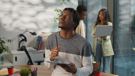 Thoughtful pondering African American informal business man businessman male guy startup leader writing ideas in office background of two multiethnic coworkers team discuss with laptop three managersの写真素材