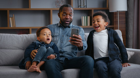 Happy African American family winning with phone at home father with two sons boys children little kids win mobile casino online victory achievement triumph celebrating smartphone joy triumph rejoiceの写真素材