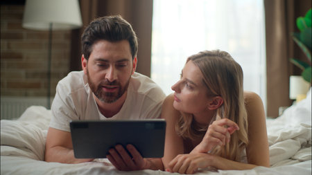 Relaxed family Caucasian couple talking pair using tablet together in bedroom man holding gadget wife woman purchasing delivery online husband play virtual game social media morning cyberspace web bedの写真素材