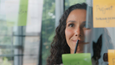 Thoughtful pensive business woman writing on sticky notes Hispanic businesswoman in company office write ideas thinking pondering with stickers project female girl organizing tasks developing on glassの写真素材