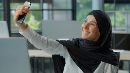 Smiling happy Arabian Muslim business woman in hijab take selfie photo with phone in office Islamic businesswoman female girl posing at workplace photographing with smartphone mobile social media vlogの写真素材