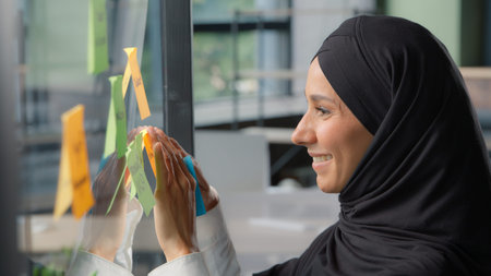 Smiling Muslim business woman stick sticky notes on glass in modern office Arabian businesswoman in...