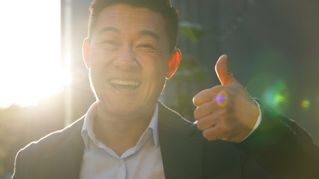 Portrait happy smiling Asian business man showing thumb up like gesture Korean businessman in city sunny sun shining male Chinese manager Japanese professional smile looking at camera agree recommendの写真素材