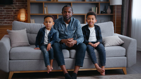 Happy African American family portrait at home living room smiling spending time in living room father dad with two boys kids sons children cuddling looking to camera on couchの写真素材