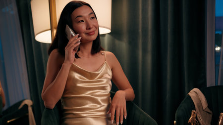 Smiling beautiful attractive Asian woman gorgeous Chinese Japanese lady Korean ethnic female at home wearing luxury dress talking pleasant conversation mobile phone call talk communicate smartphoneの写真素材
