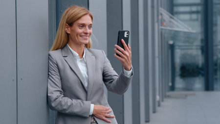 Caucasian middle-aged business woman talking phone video call in city leaning on wall of office building adult businesswoman female chatting online conference online webcam mobile remote conversationの写真素材