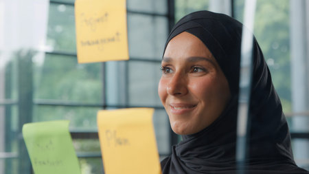 Satisfied business woman analyzing stickers on glass in office Muslim businesswoman in hijab CEO...