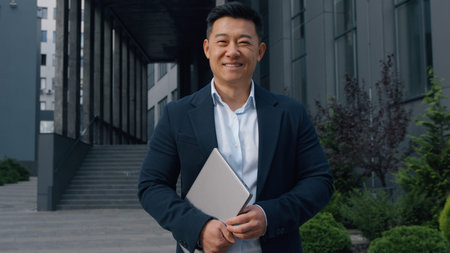 Asian middle-aged businessman with laptop smiling Korean business man posing in city holding...
