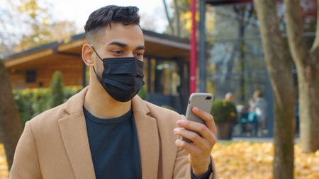 Hispanic man in face medical mask looking in mobile phone using app smartphone in park autumn fall season Arabian guy male chatting telephone social media chat outdoors positive online communicationの写真素材