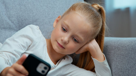 Relaxed Caucasian child little girl daughter kid teen with phone resting on couch in living room using mobile application modern technology online chat internet games at home with smartphone deviceの写真素材