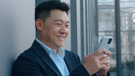 Smiling Asian middle-aged business man looking in phone worker businessman smile positive carefree use mobile application in city leaning building wall happy chattingの写真素材