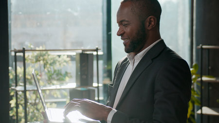 Smiling ethnic man African American businessman business agent with laptop in corporate office adult male executive professional working online working digital data analysis review browsing computerの写真素材