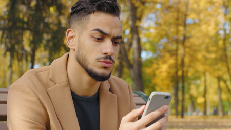 Portrait serious frown Hispanic man using mobile phone guy use app in autumn park reading bad news chat pensive negative emotion relaxing on bench in city looking at smartphone telephone concentratedの写真素材