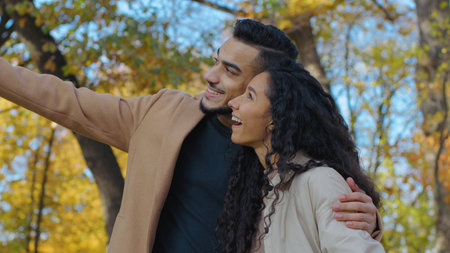 Smiling Hispanic couple smiling talking on date in autumn park talk looking on gold leaf foliage fall season in city happy woman with man hugging cuddling pointing on yellow trees loveの写真素材