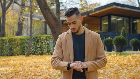 Worried Hispanic man guy in autumn park look around waiting for meeting looking at wrist watch disappointed nervous wait sad male standing outdoors in fall season lateness problem cancel appointmentの写真素材