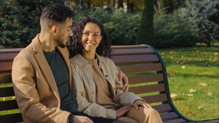 Cheerful hispanic couple in autumn park talking happy relaxing on bench in city communicate smiling Arabian man with lovely woman talk hugging love weekend fall season outdoors pointing away discussの写真素材