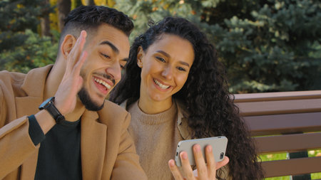 Happy couple talking on video call with mobile phone in autumn city park sitting on bench. Woman and man online talk on conference with smartphone greeting chat.の写真素材