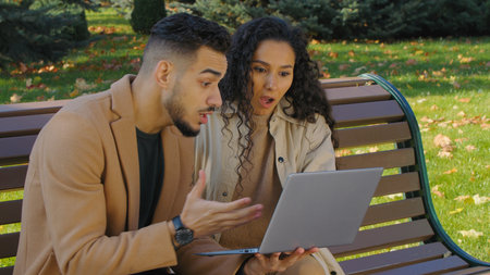 Happy emotional hispanic couple winning with laptop in city autumn park sitting on bench family arabian man caucasian woman multiracial married girl and guy with computer win achievementの写真素材