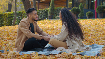 Happy Hispanic couple on date in autumn park talking laughing having fun sitting on blanket on yellow foliage woman in love talk smile laugh holding hands outdoors affection weekend natureの写真素材