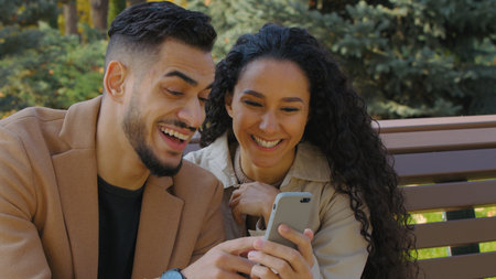 Hispanic happy couple guy with girl in autumn park on bench looking in phone cheerful family man woman discussing shopping online booking restaurant date talking smiling look in mobile app smartphoneの写真素材