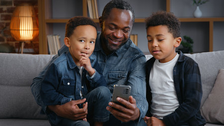 Happy African American family looking in smartphone at home father with two little boys children kids enjoying mobile phone app social media internet smiling watching funny video on couch talkingの写真素材