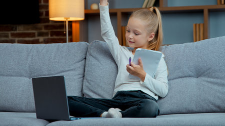 Caucasian child girl kid daughter pupil listen online lesson writing raising hand answer teacher e-learning with laptop at home educational internet lesson virtual class write notes studying learningの写真素材