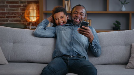 Happy African American family daddy using phone son jumping running surprise dad cuddling laughing together with mobile smartphone boy kid child at home enjoying relationship fathers day laughingの写真素材