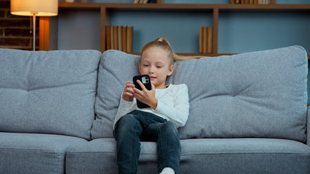 Active little child daughter kid girl jump on sofa using mobile phone apps generation falling on couch in living room scrolling smartphone social media networks internet chatの写真素材