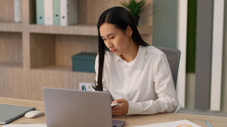 Serious attentive Asian woman female worker girl manager businesswoman secretary using laptop video call conference business meeting listening nods head writing notes at officeの写真素材