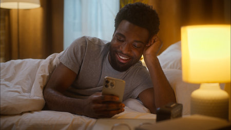 Happy African American man laughing use mobile phone bedroom evening ethnic guy male client customer scrolling social media before sleep watching funny video enjoy smartphone relax at home bed nightの写真素材