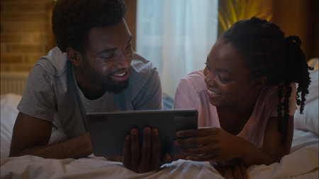 Cute happy family romantic laughing African American couple girlfriend boyfriend lying on bed together holding tablet device gadget look screen watching film movie cuddle at home bedroom late eveningの写真素材