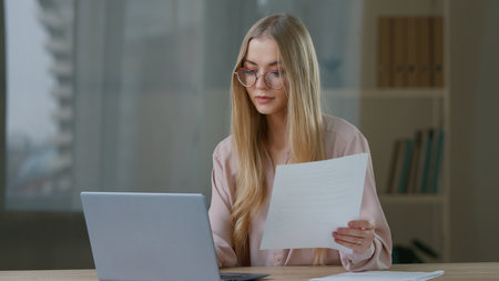 Caucasian concentrated business woman working with laptop checking papers in office focused businesswoman girl female entrepreneur indoors reading documents data review working online specialist workの写真素材