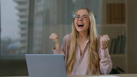 Happy Caucasian business woman winning with laptop in office winner champion success businesswoman female employer girl celebrating win with computer work job offer growth victory yelling achievementの写真素材