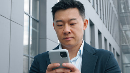 Pensive Asian Korean business man with mobile phone thinking typing message middle-aged Korean businessman in city deciding think idea browsing chatting smartphone app Japanese Chinese male workerの写真素材