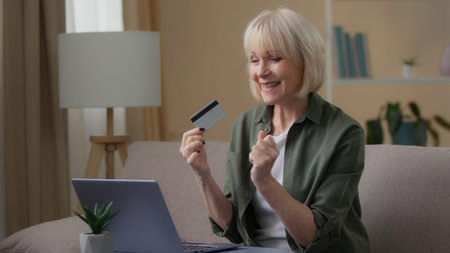 Elderly Caucasian female old 60s woman buying online with laptop and credit card at home smiling...