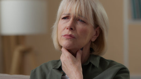 Old Caucasian woman feeling pain in throat at home holding neck retired sick female senior lady feel...