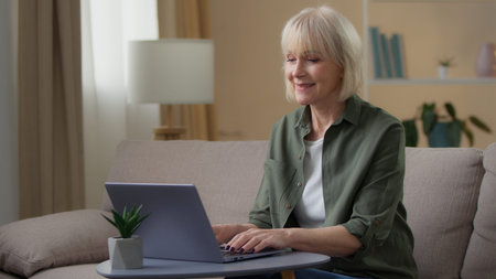 Old Caucasian woman smiling looking in laptop using cyberspace modern technology at home happy...
