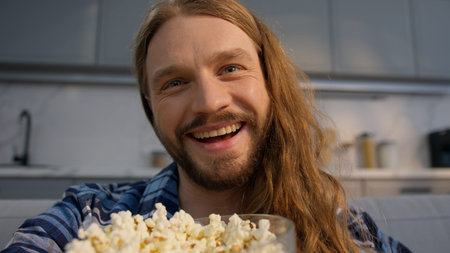 Laughing smiling Caucasian man watching TV at home eating popcorn guy male enjoying funny humorous online show television program movie film joke fun carefree positive relaxed indoors entertainmentの写真素材