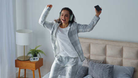 Woman jumping dancing on bed listening music in earphones smiling moving hands party jump in bedroom home female girl listen headphones audio song sound holding mobile phone delightの写真素材