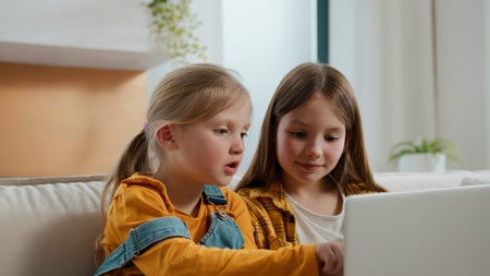 Two little Caucasian girls talking communicate conversation looking in laptop pointing in screen kids daughters children siblings sisters at home couch talk enjoying computer technology playing gameの写真素材