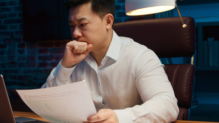 Overload Asian tired Korean business man with laptop reading documents at evening office middle-aged businessman working online bankruptcy problem insomnia headache at nightの写真素材