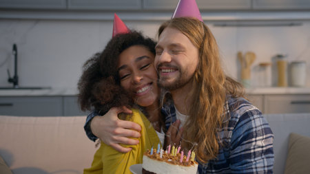 Joyful multiracial couple celebrating birthday at home Caucasian man holding cake African American woman blowing candles smiling hugging celebration boyfriend girlfriend family surprise party togetherの写真素材