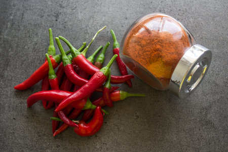 Red chili peppers and paprika powder in glass spice jar on stone table. Dark grey background with...