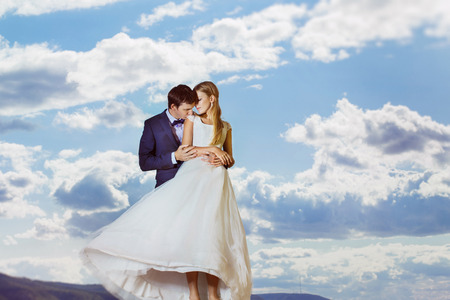 A beautiful young wedding couple is embracing at the blue summer sky with clouds background.の写真素材